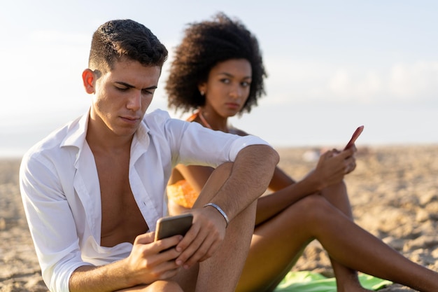 Een meisje van Afrikaanse afkomst is jaloers en kijkt toe hoe haar vriend zijn gsm-berichten verbergt, terwijl ze op vakantie zijn, zittend op het strand.