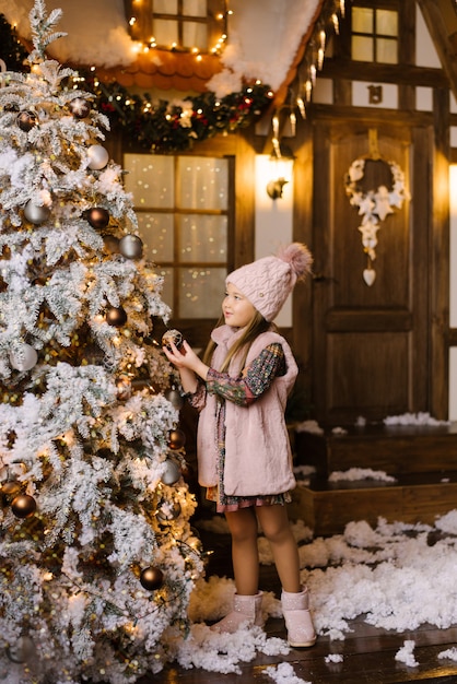 Een meisje van vijf of zes jaar in winter roze kleding en ugg laarzen staat in de buurt van de kerstboom