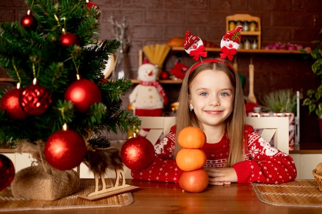 Een meisje verwent zich met mandarijnen en onderscheidt zich van hen een sneeuwpop in een donkere keuken bij een kerstboom met rode ballen, het concept van nieuwjaar en Kerstmis