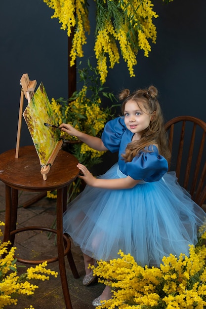 Een meisje zit in de buurt van een mimosa en schildert een foto in een blauwe jurk mimosa borstel boeket meisje