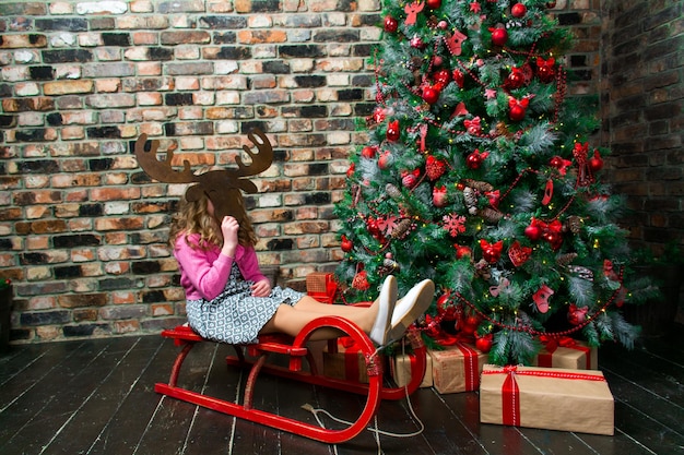 Een meisje zit op een slee bij de kerstboom in een masker van een kersthert Merry Christmas