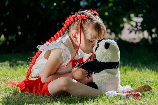 Een meisje zit op het gazon met een speelgoedpanda en kust hem op de neus. Hoge kwaliteit foto