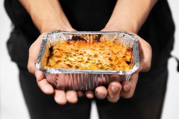 Een menselijke hand die pasta macaroni schotel vasthoudt in een aluminiumfolie beker met een witte achtergrond