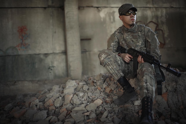 Een militair in een grijs camouflage-uniform met een pet en een tactische bril met een grote zwarte atoom in zijn handen zit op de grond in een groot verwoest gebouw