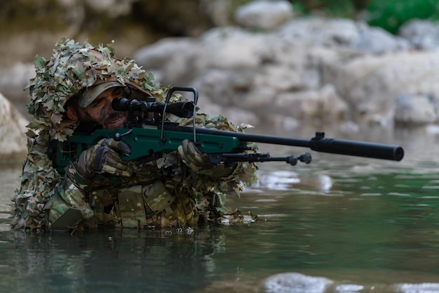 Een militair of airsoftspeler in een camouflagepak sluipt over de rivier en richt met een sluipschuttersgeweer opzij of op doel.