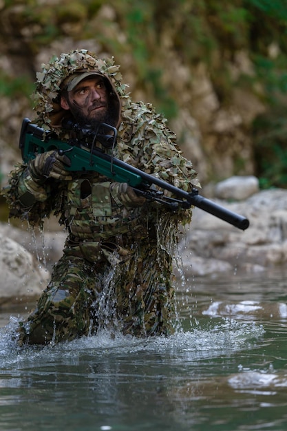 Een militair of airsoftspeler in een camouflagepak sluipt over de rivier en richt met een sluipschuttersgeweer opzij of op doel.