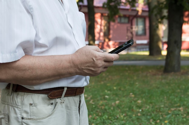 Een mobiele telefoon in de grote handen van een oudere man in een wit overhemd tijdens een wandeling in het park