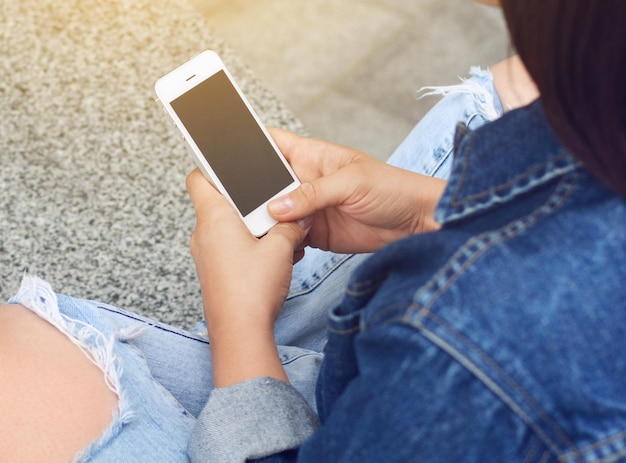 Een mockup-afbeelding van handen met een witte mobiele telefoon met een leeg zwart scherm Een jong hipstermeisje gebruikt een smartphone terwijl ze zit Conceptuele foto