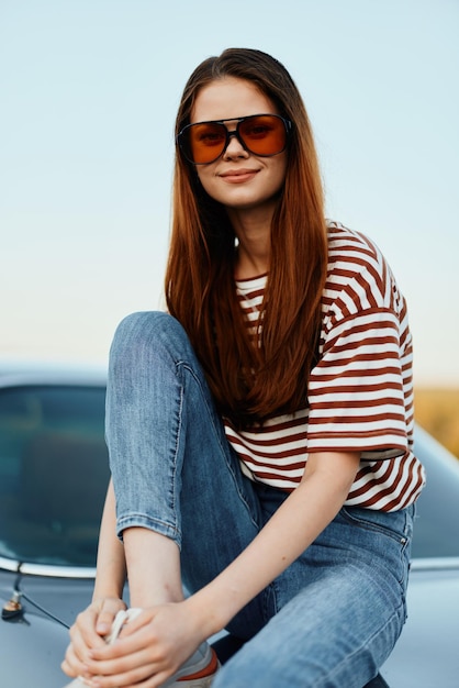 Een modevrouw in een stijlvolle zonnebril, een gestreept t-shirt en een spijkerbroek zit op de kofferbak van een auto en kijkt naar de prachtige natuur van de herfst Reislevensstijl