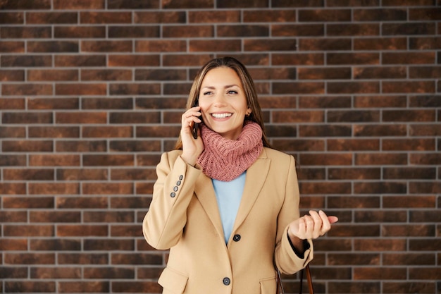 Een modieuze zakenvrouw die voor een bakstenen muur staat en aan de telefoon praat