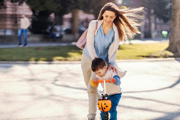 Een moeder duwt zoon op een scooter in een park