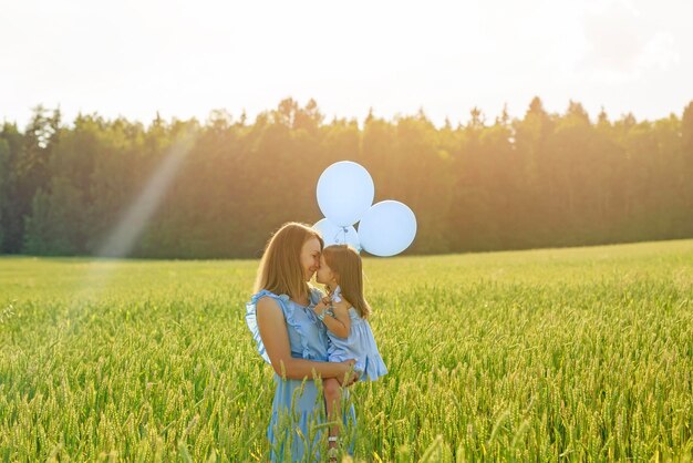 Een moeder en dochter in een groen tarweveld met blauwe ballonnen omhelzen elkaar teder in liefde