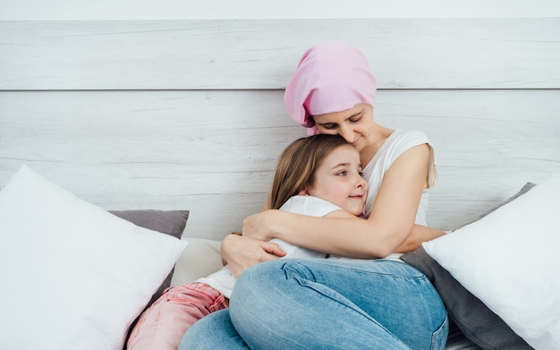 Een moeder met kanker draagt een roze hoofddoek en omhelst teder haar mooie blondharige dochter. Ze zitten allebei op het bed met een witte achtergrond