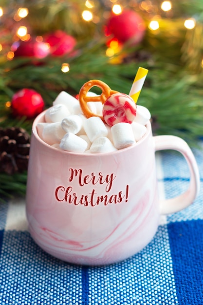 Een mok met de tekst merry christmas met marshmallow op een achtergrond van een tak van een kerstboom