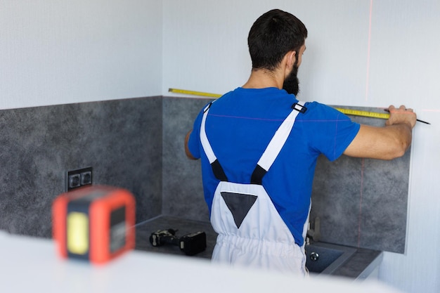 Een monteur gebruikt een meetband en een laserniveau om wandpanelen en kasten in keukens te installeren