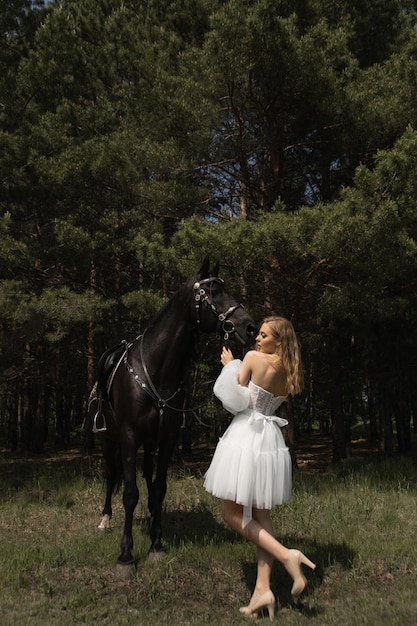 Foto een mooi blank meisje in een trouwjurk staat naast een paard in het bos