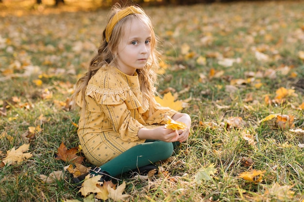 Een mooi blond meisje in een gele jurk zit in een herfstpark met esdoornbladeren