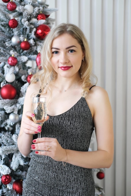 Een mooi blond meisje in een grijze glanzende jurk staat tegen de achtergrond van een kerstboom met een glas champagne in haar handen