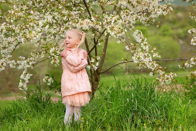 Een mooi blond meisje in een roze outfit loopt in de tuin naast een bloeiende kersenboom op de zonnige lente dag.