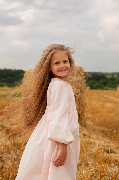 een mooi blond meisje met lang haar in een linnen jurk staat bij een hooiberg