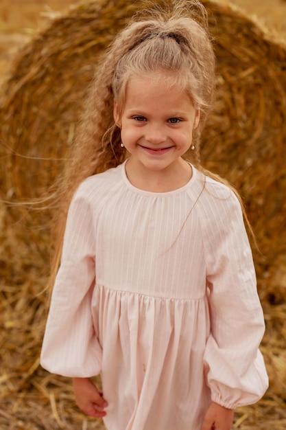 een mooi blond meisje met lang haar in een linnen jurk staat bij een hooiberg