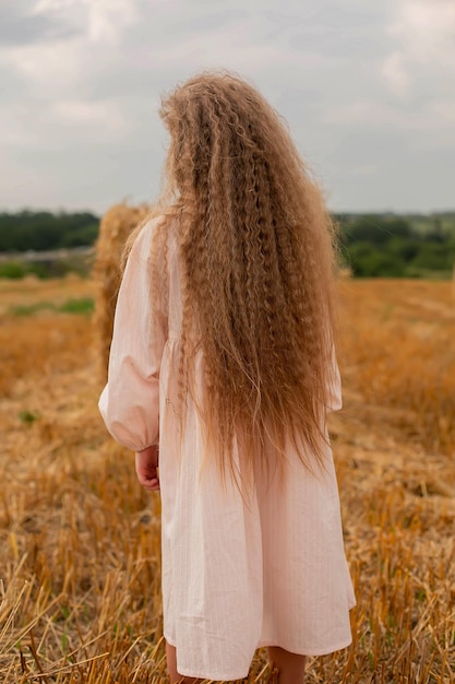 een mooi blond meisje met lang haar in een linnen jurk staat met haar rug op een gemaaid veld