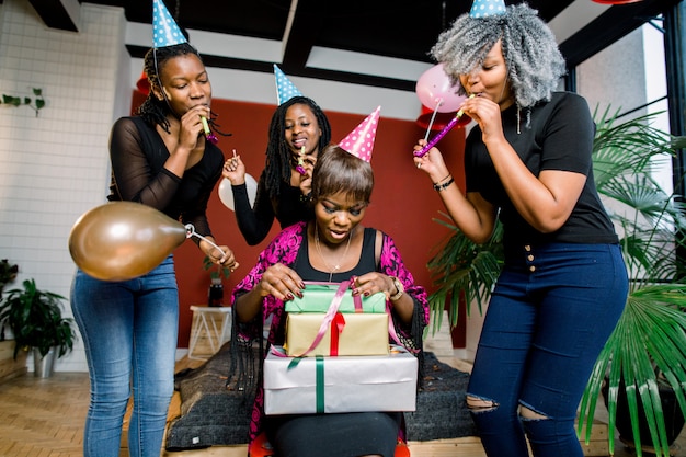 Een mooi glimlachend Afrikaans meisje opent een cadeau op haar verjaardagsfeestje. Gelukkige Afrikaanse meisjes in feestmutsen en met blazende hoorns staan rond feestvarken en glimlachen