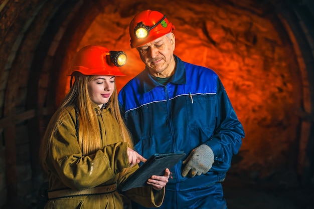 Een mooi jong meisje in een rode helm en met een tablet in haar handen bevindt zich met een mijnwerker in een kolenmijn.