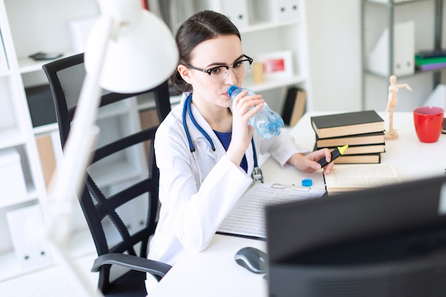 Een mooi jong meisje in een witte robe zit achter een computerbureau, drinkt water en kijkt naar de monitor.