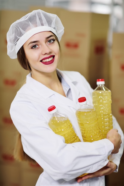 Een mooi jong meisje in wit uniform of een werkende badjas met een fles zonnebloemolie in haar handen op het oppervlak van de voedselproductielijn van geraffineerde olie.