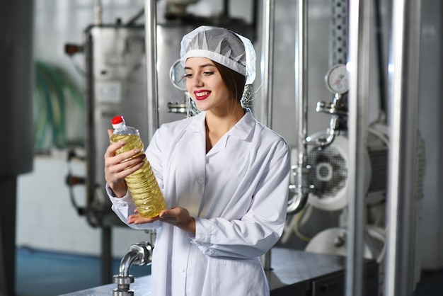 Een mooi jong meisje in wit uniform of een werkende badjas met een fles zonnebloemolie in haar handen op het oppervlak van de voedselproductielijn van geraffineerde olie.