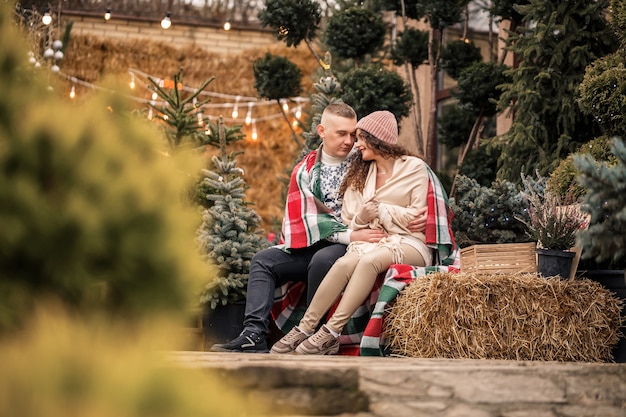 Een mooi jong stel in witte kleren zit bij een kerstboom in de tuin. Gelukkig man en vrouw, romantiek, kerstviering, plezier, liefde.