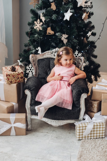 Een mooi Kaukasisch meisje met kort blond golvend haar in een schattige roze jurk verheugt zich in de buurt van de kerstboom en veel cadeautjes in een grote lichte kamer van het huis