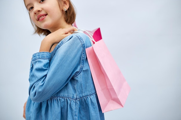 Een mooi kindmeisje in een casual spijkerjurk houdt roze boodschappentassen in de hand