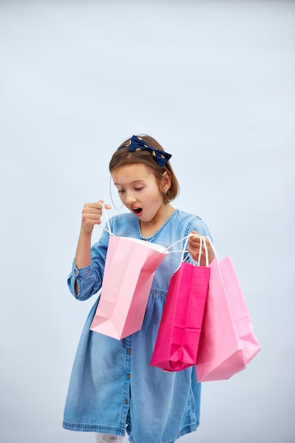 Een mooi kindmeisje in een casual spijkerjurk houdt roze boodschappentassen in de hand