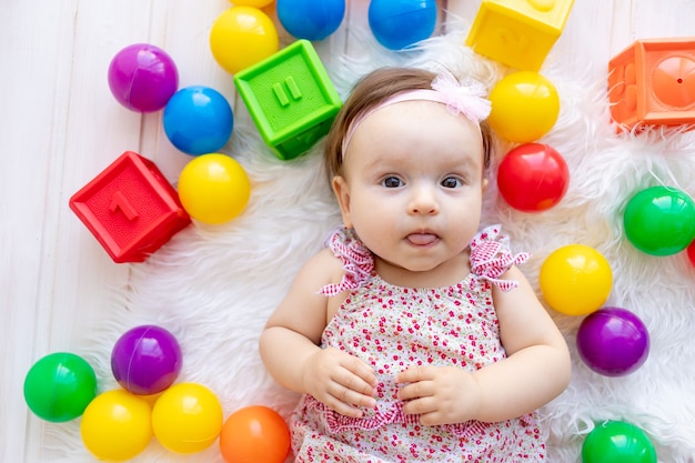 Een mooi klein babymeisje ligt in rode kleren op een witte Mat onder speelgoedballen en kubussen