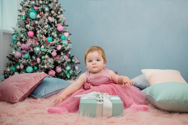 Een mooi klein meisje in een tutu-jurk zit op het bed met een cadeau onder de kerstboom