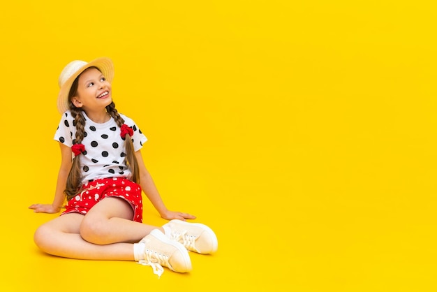 Een mooi klein meisje in korte broek met stippen en een strohoed wijst naar uw advertentie zomervakantie een tienermeisje zit op een gele geïsoleerde achtergrond