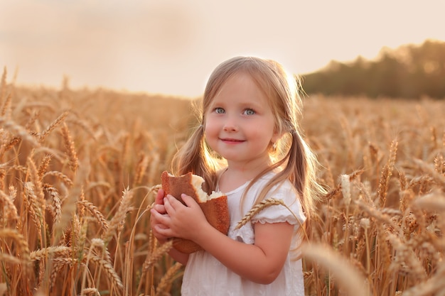 een mooi klein meisje in witte kleren blonde staat in een tarweveld met brood