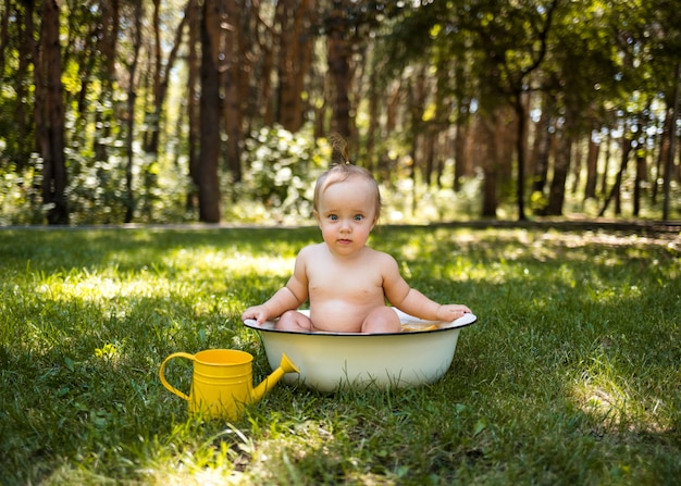 Een mooi klein meisje zit in een bad met water en een gieter en kijkt naar de camera op het groene gras