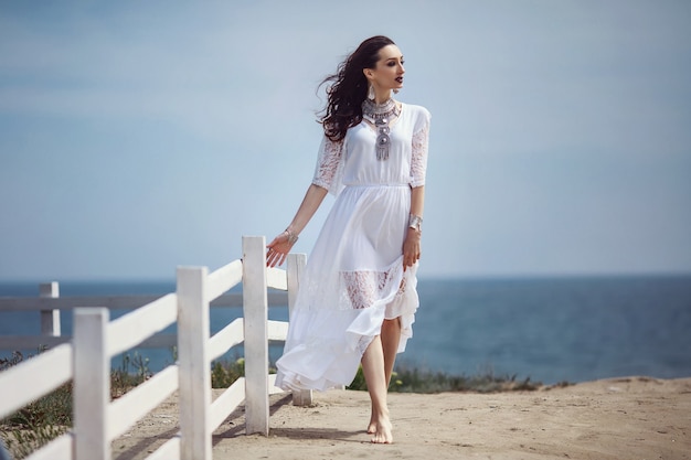 Een mooi meisje, bruid, in een witte jurk, op blote voeten, lopend bij een wit hek