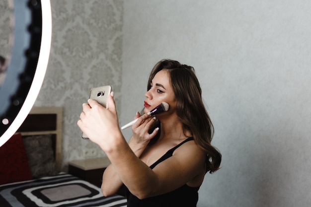 Een mooi meisje doet make-up op haar gezicht met een penseel en maakt foto's van zichzelf aan de telefoon.