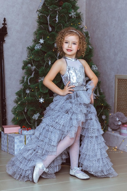 Een mooi meisje in een elegante zilveren jurk staat bij de kerstboom