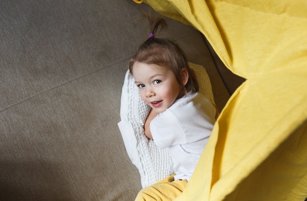 Een mooi meisje in een gele broek en een wit T-shirt lacht en speelt in de tipi thuis op de bank. Home games voor kinderen