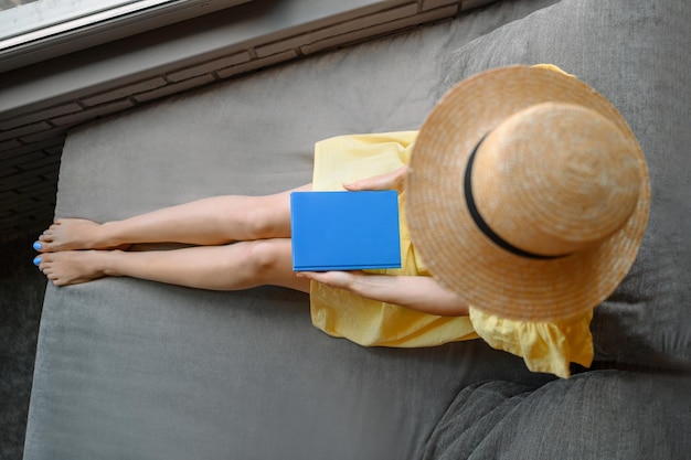 Een mooi meisje in een gele zomerjurk en een strohoed zit met een boek op het balkon