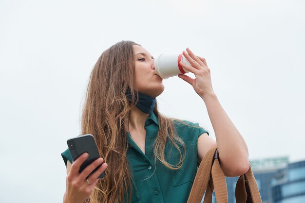 Een mooi meisje in een leeggelopen gezichtsmasker met lang haar drinkt koffie en houdt een smartphone in haar hand in het centrum