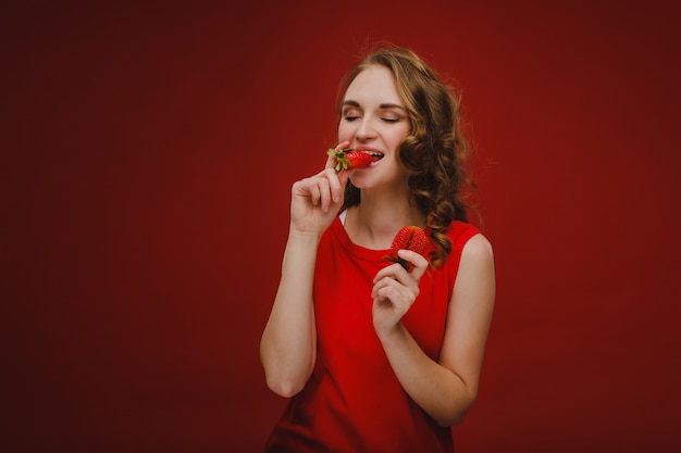 Een mooi meisje in een rode jurk op een rode achtergrond houdt een aardbei in haar handen en glimlacht