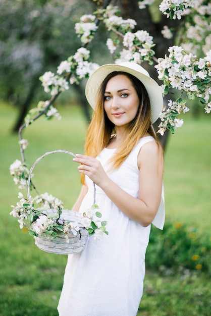 Een mooi meisje in een witte jurk en een witte hoed met een rand met een mand met takken van een bloeiende appelboom staat in de lentetuin