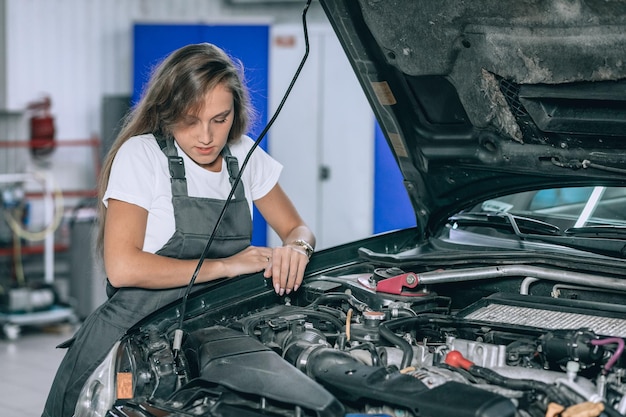 Een mooi meisje in een zwarte jumpsuit en een wit t-shirt lacht en controleert het oliepeil in een zwarte auto in de garage.