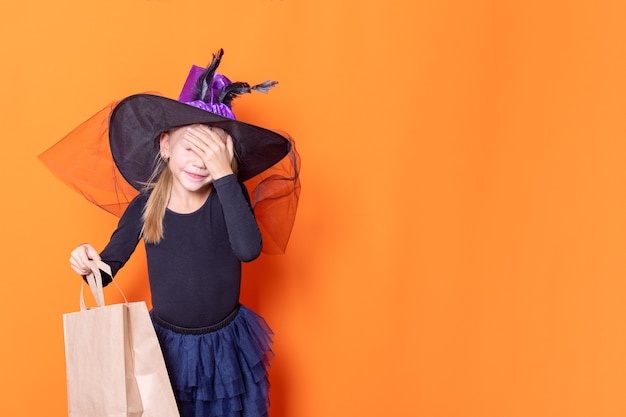 Een mooi meisje in een zwarte jurk en een heksenhoed houdt een ambachtelijke papieren zak vast en bedekt haar gezicht met haar hand, zich afvragend, op een oranje achtergrond. Het concept van geweldige geschenken. Halloween winkelconcept